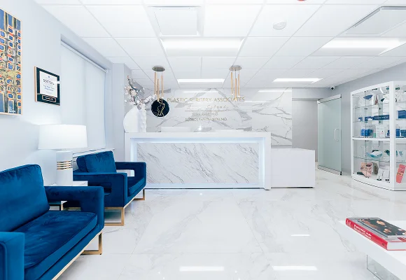A bright, clean view of the office reception area with white marble floors, two teal velvet chairs, and a glass display case of medical products.
