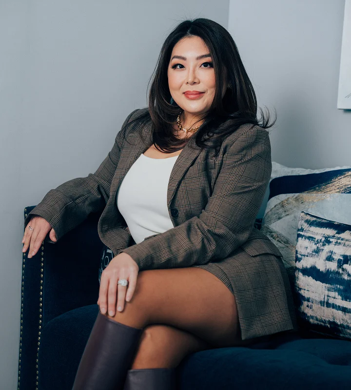 A professional portrait of Dr. Rhee, wearing a brown plaid blazer and white top, sitting confidently on a navy blue sofa.