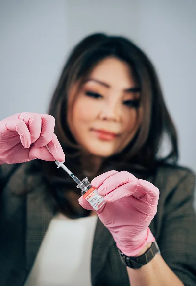A close-up of Dr. Rhee wearing pink medical gloves, carefully drawing liquid from a small glass vial into a syringe.