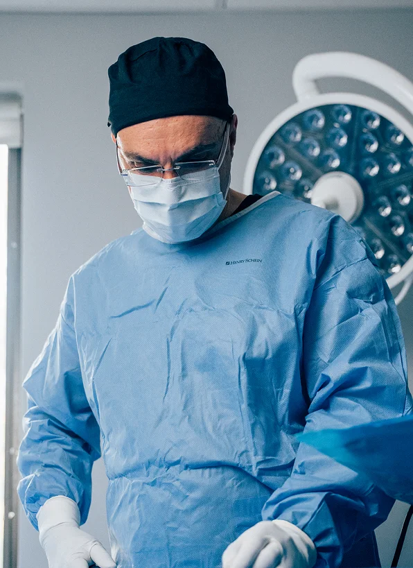 Dr. Papafragkou, wearing a blue surgical gown, a black scrub cap, a medical mask, and glasses, looks downward with focus while performing a procedure.