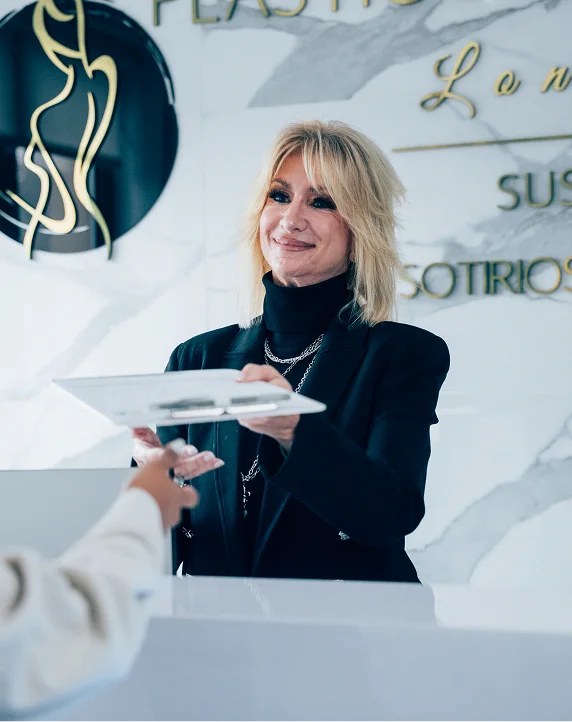 A blonde woman in a black blazer smiles warmly as she hands a white clipboard over a reception desk to a patient.