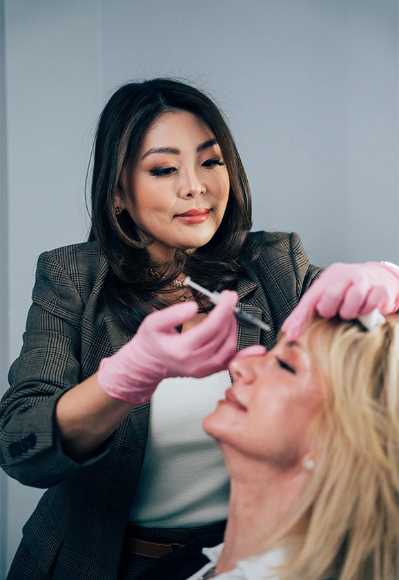 Dr. Rhee wearing a plaid blazer and pink gloves administers an injection to a patient's forehead.