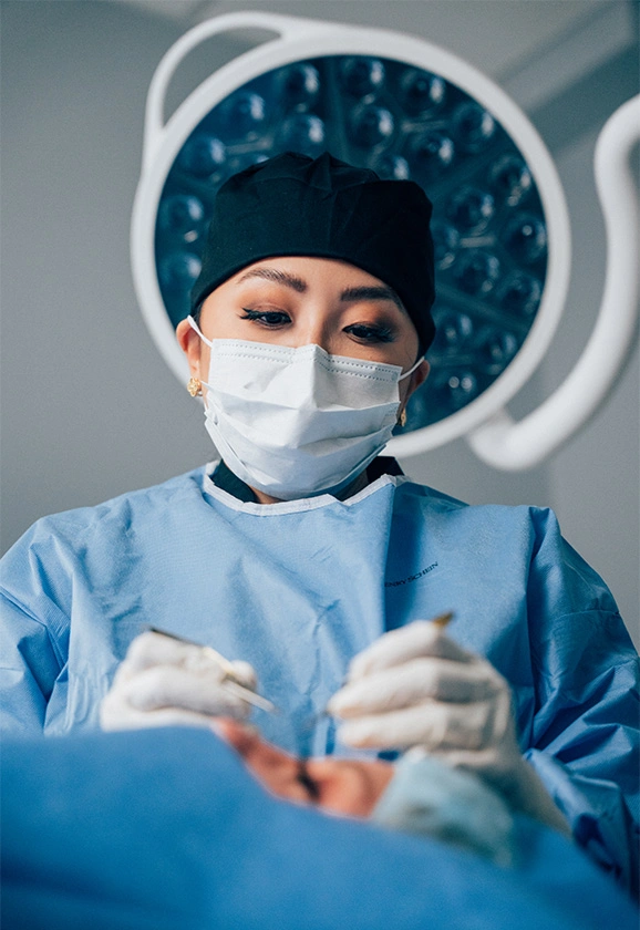Dr. Rhee wearing a surgical mask and cap, looking down while performing a procedure under bright surgical lights.