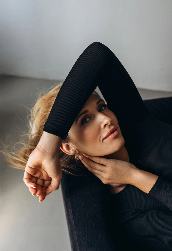 A woman with blonde hair wearing a black long-sleeve top reclines with her arm raised above her head.