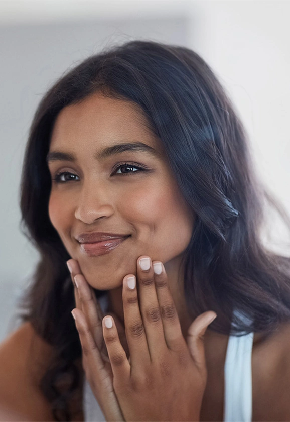 A woman with dark skin and hair smiles while gently touching her face with her fingers.