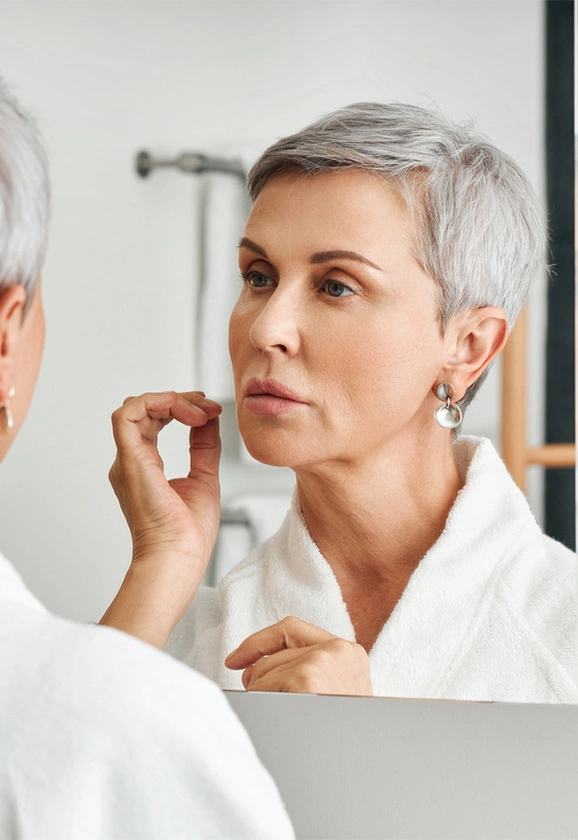 A woman with short grey hair looks at her reflection in a mirror while wearing a white bathrobe.