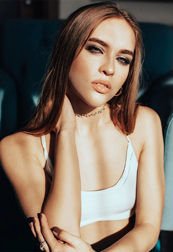 A woman with long brown hair and dramatic eye makeup poses in a white tank top and gold chain necklace.