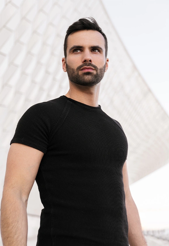 A man with a beard wearing a black ribbed t-shirt looks off to the side against a modern white architectural background.