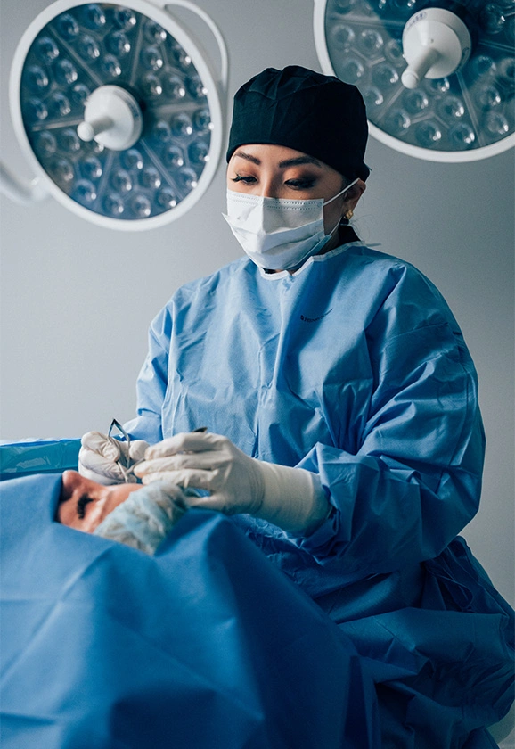Dr. Rhee, wearing a surgical mask and cap, performs a meticulous procedure under bright operating room lights.
