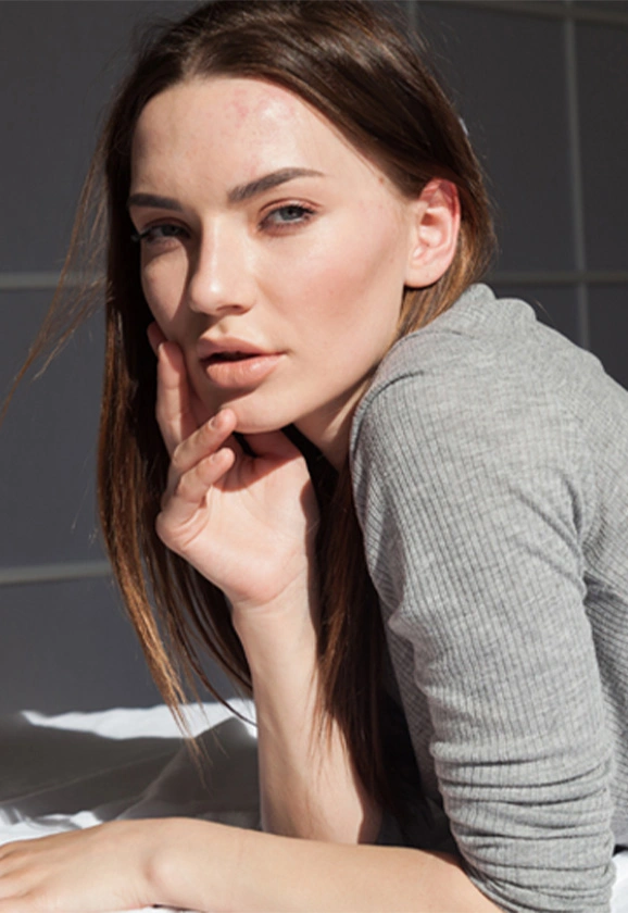 A woman with long dark hair rests her chin on her hand, looking directly at the camera in bright natural light.