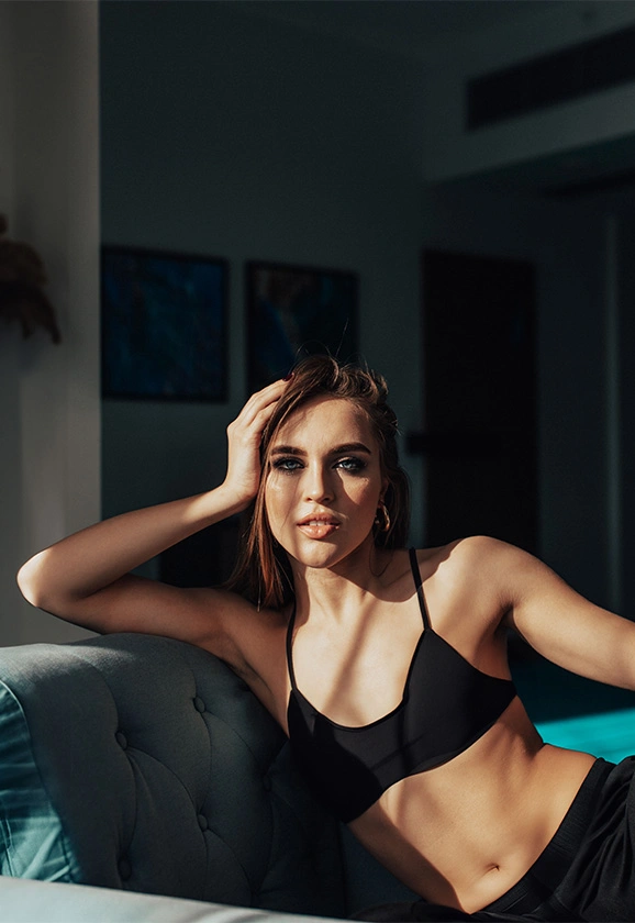 A woman in a black athletic top reclines on a blue velvet sofa in a sunlit room.