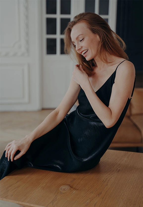 A woman with long strawberry-blonde hair wearing a black slip dress sits on a wooden table, smiling.