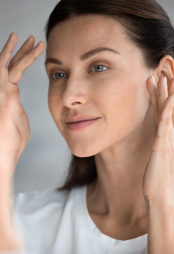 A woman with dark hair pulled back, gently touching her face while looking toward her reflection.