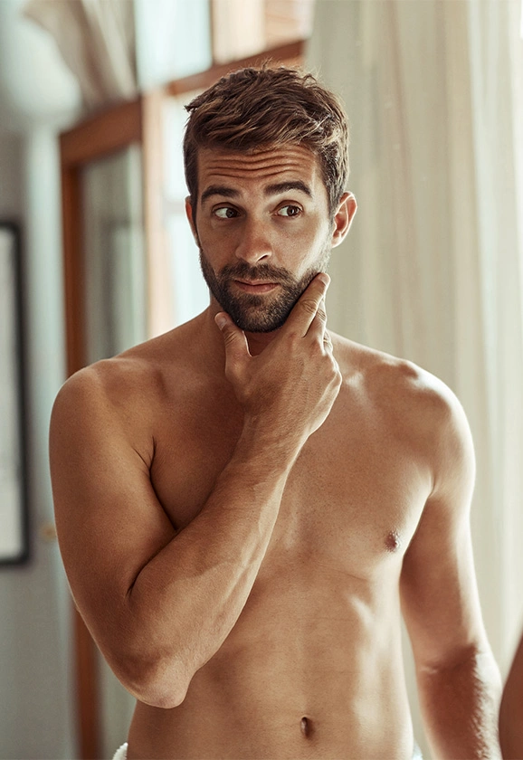 A man with a beard stands shirtless in a sunlit room, looking off to the side with one hand on his chin.