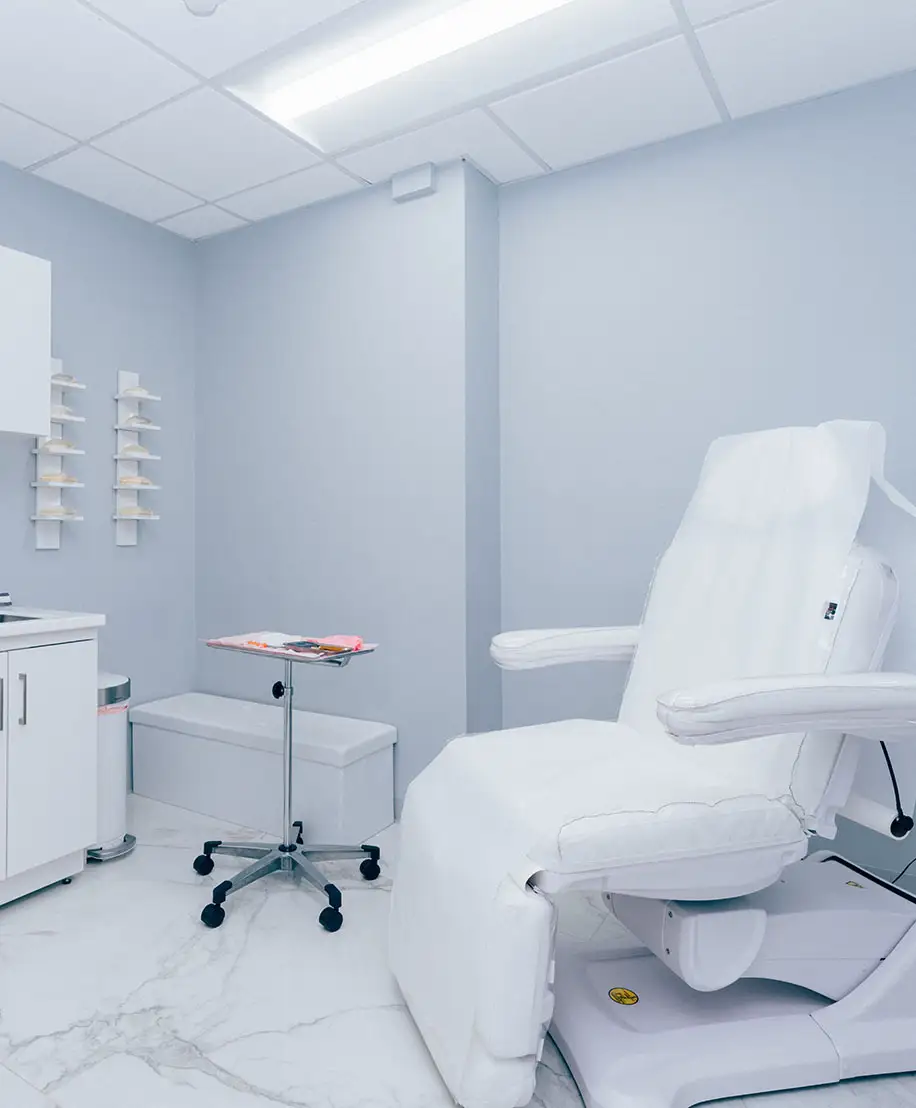 An interior view of a bright, modern medical treatment room with a white adjustable procedure chair and sterile equipment.