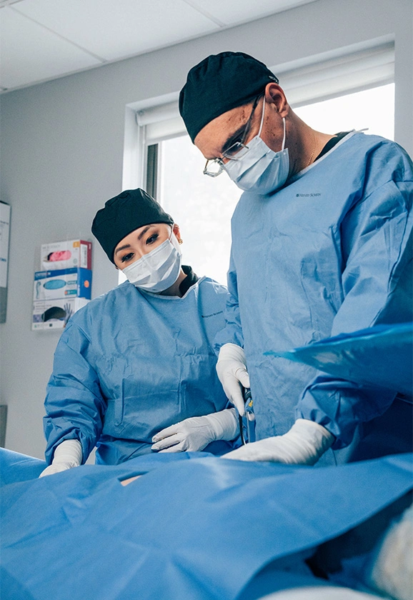 Dr. Rhee and Dr. Papafragkou working together in the operating room, wearing blue surgical gowns and masks.