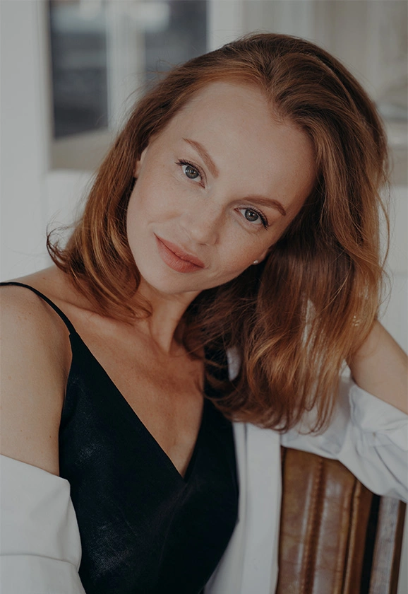 A woman with reddish-brown hair and a black top smiles softly while leaning forward.